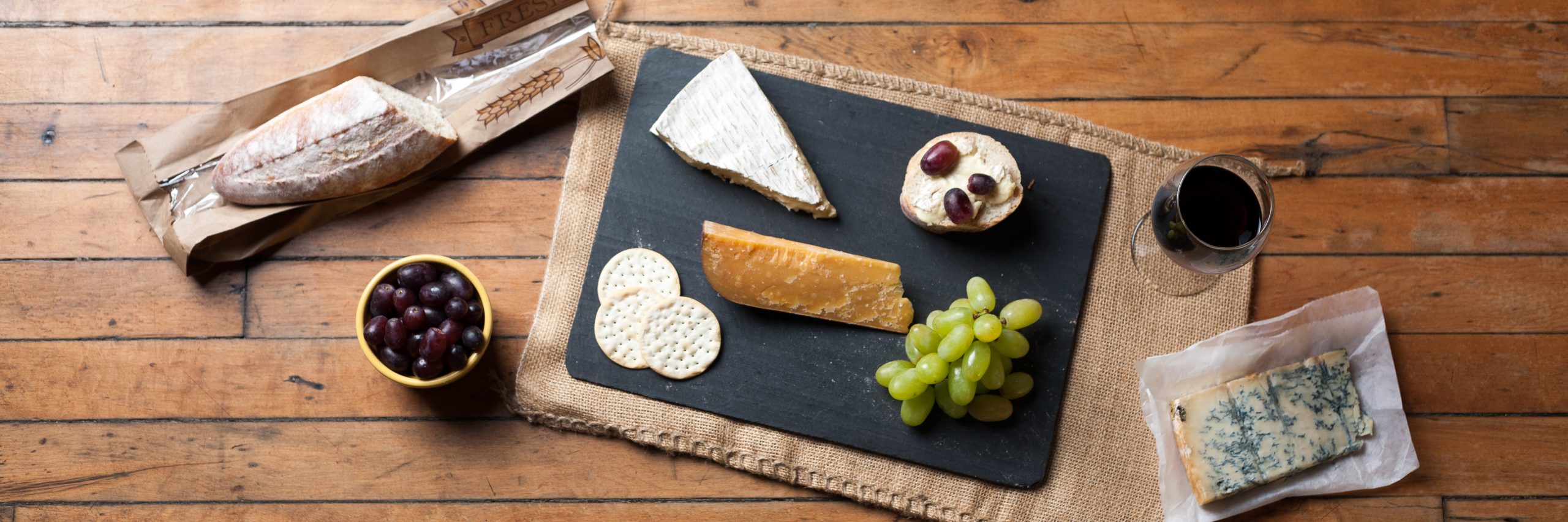 A slate of cheeses with grapes, crackers, a baguette and a glass of red wine on a wooden table