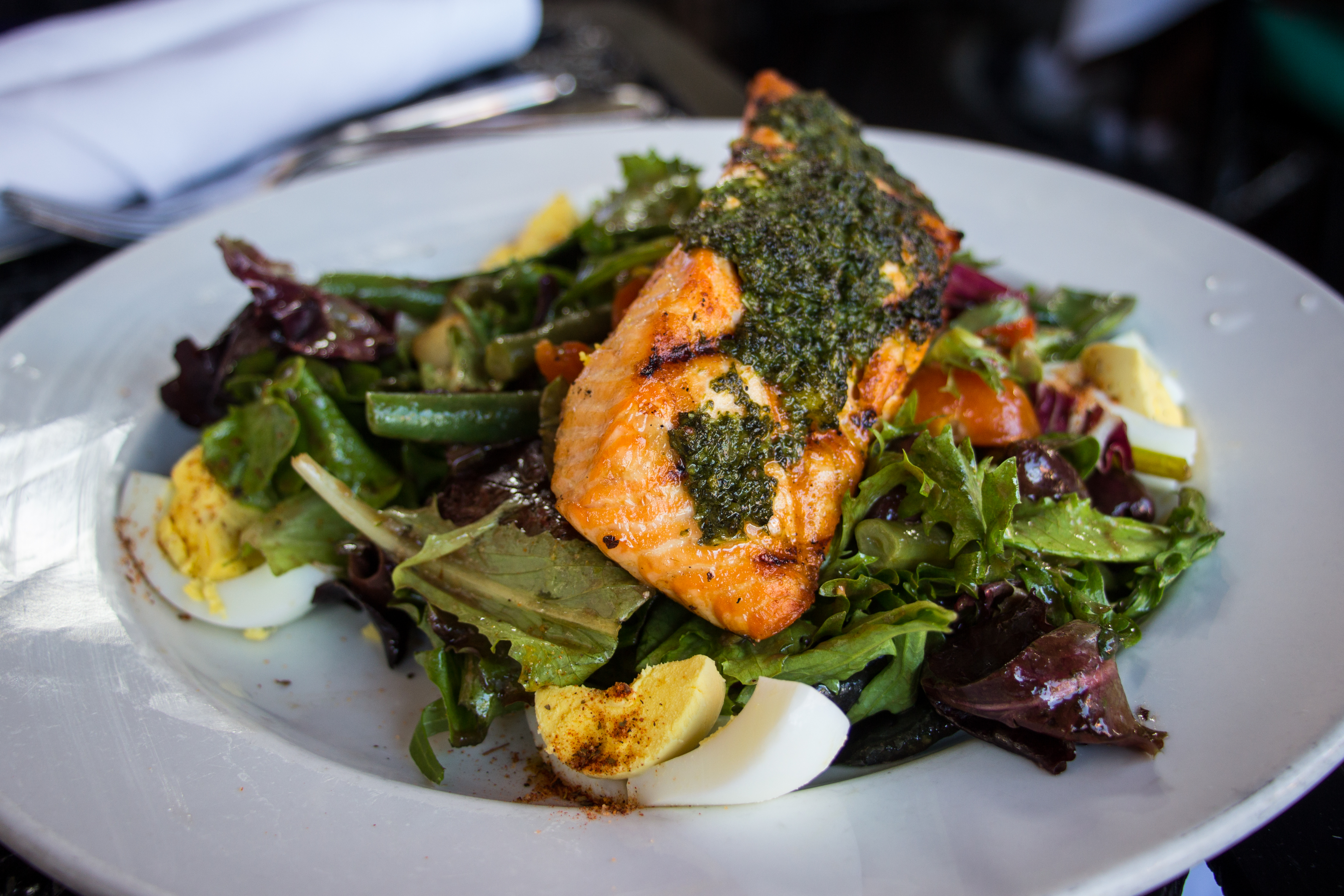 A lunch plate of grilled salmon with herb sauce on mixed leaves, green beans, olives and boiled egg