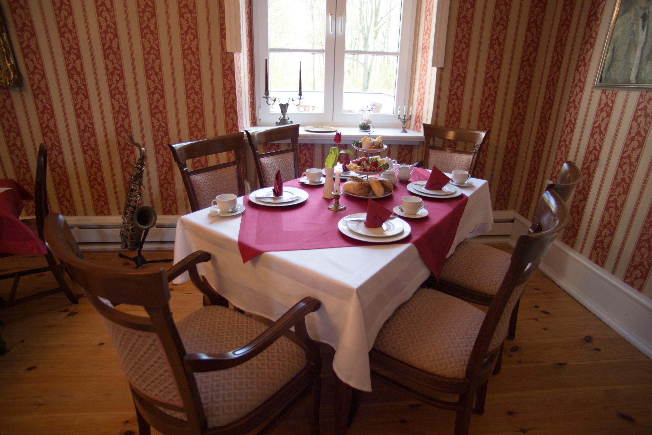 A quiet dining room with a single table laid in white and burgundy linen, cups, plates and a tiered stand of pastries