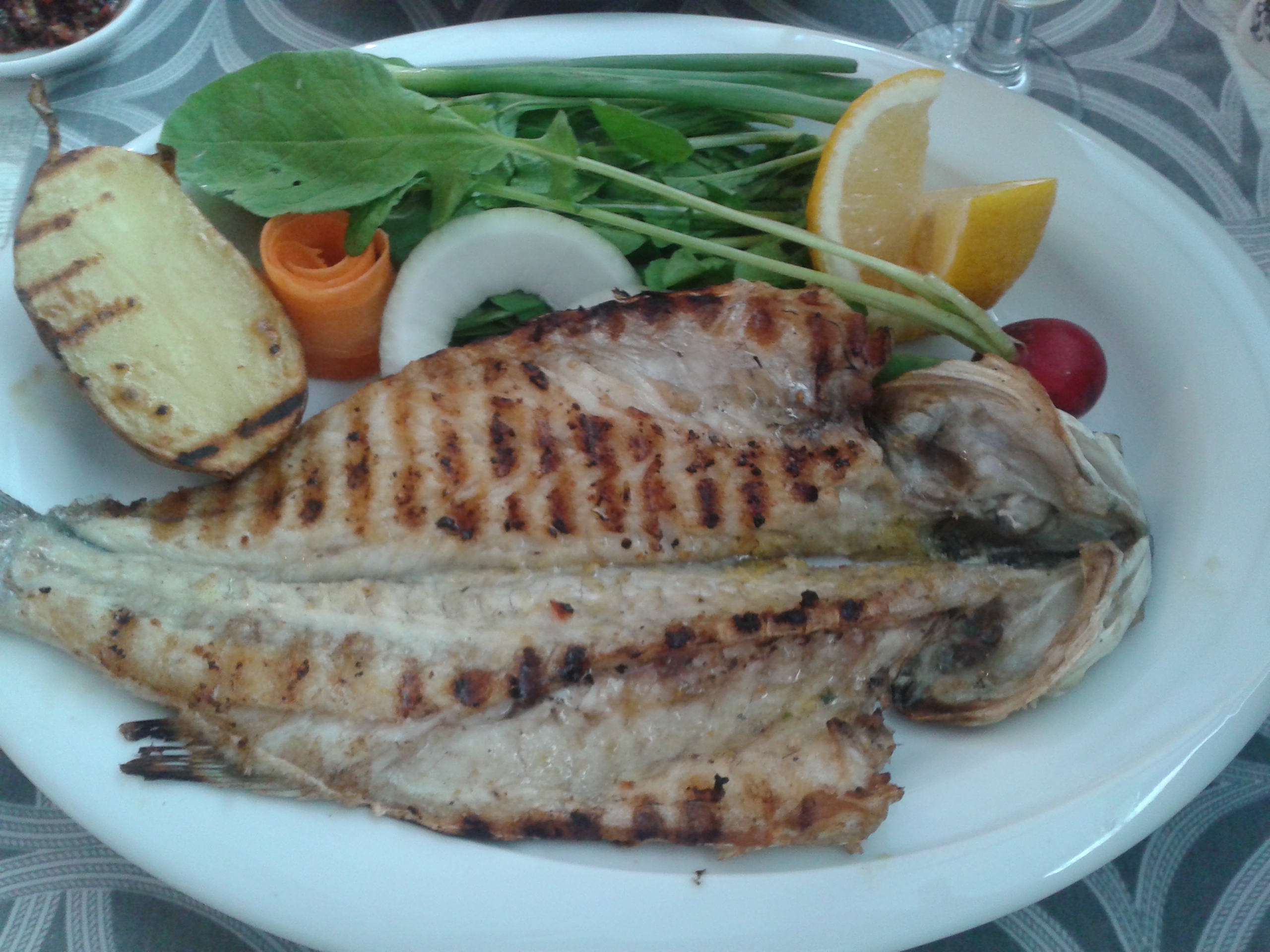 A whole grilled sea bass with a grilled potato, rocket and lemon wedges on a white plate