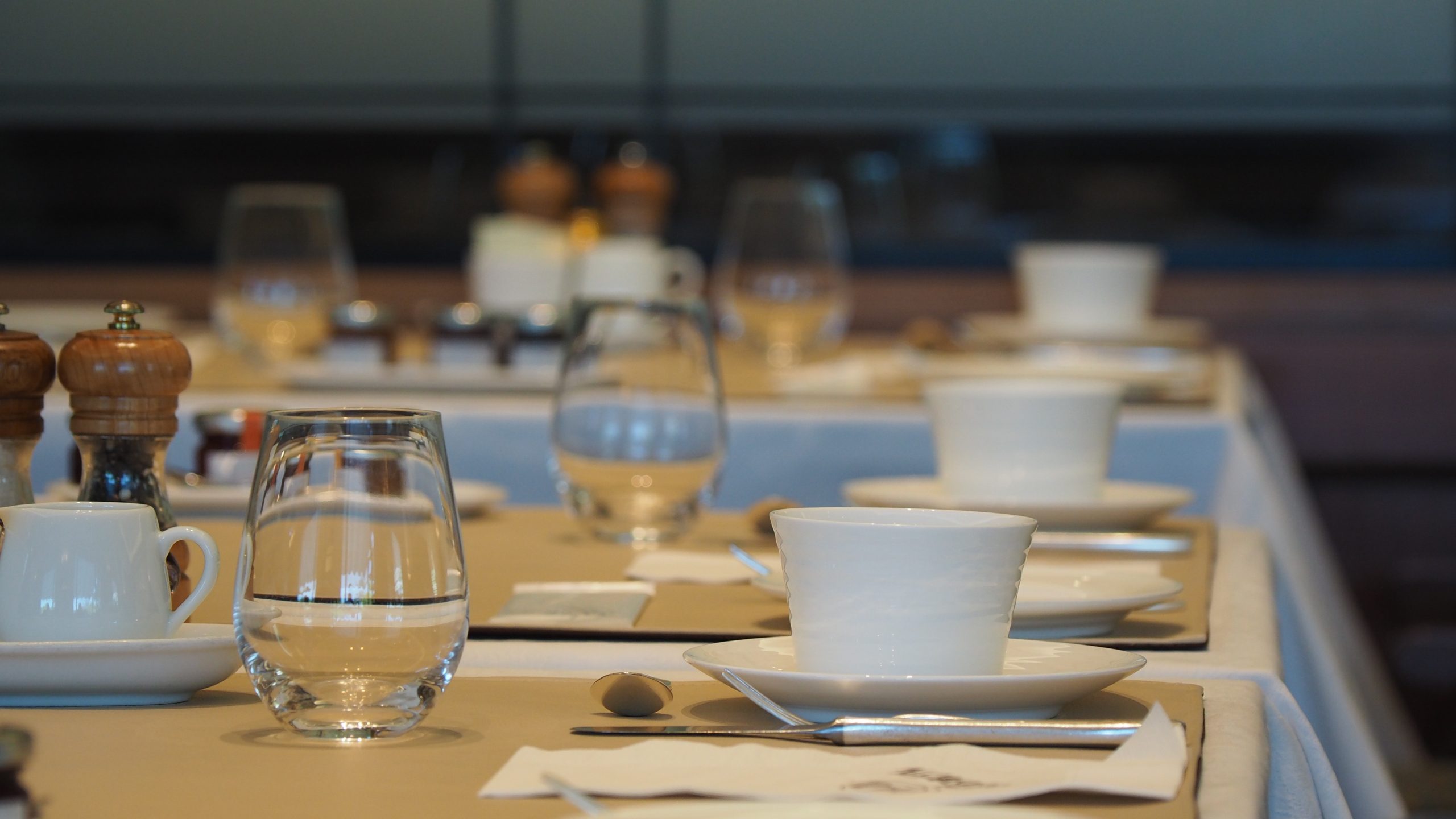 Restaurant tables laid with water glasses, white cups and saucers, cutlery and pepper mills ready for service