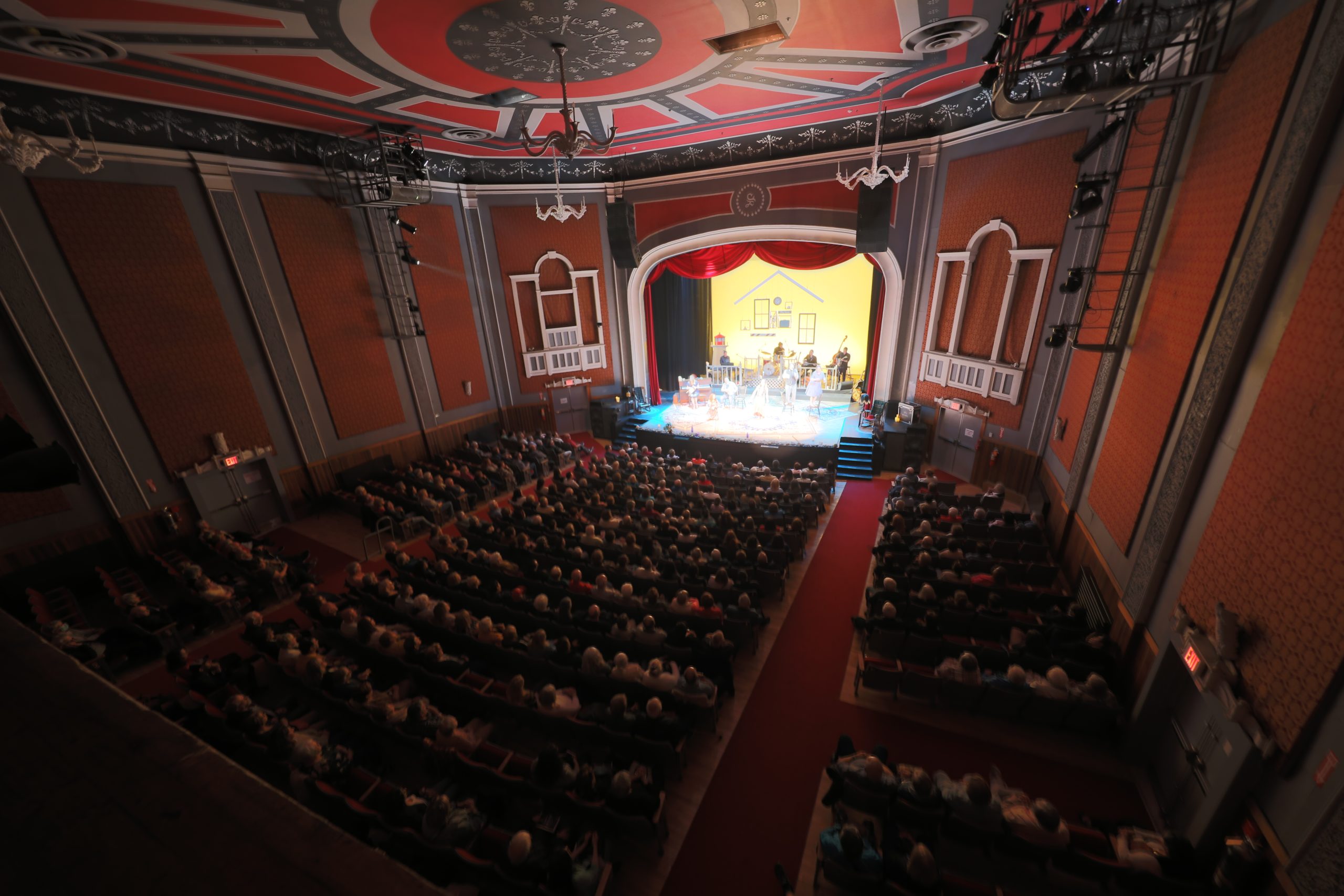 A full theatre auditorium seen from the balcony, with the audience facing a lit stage
