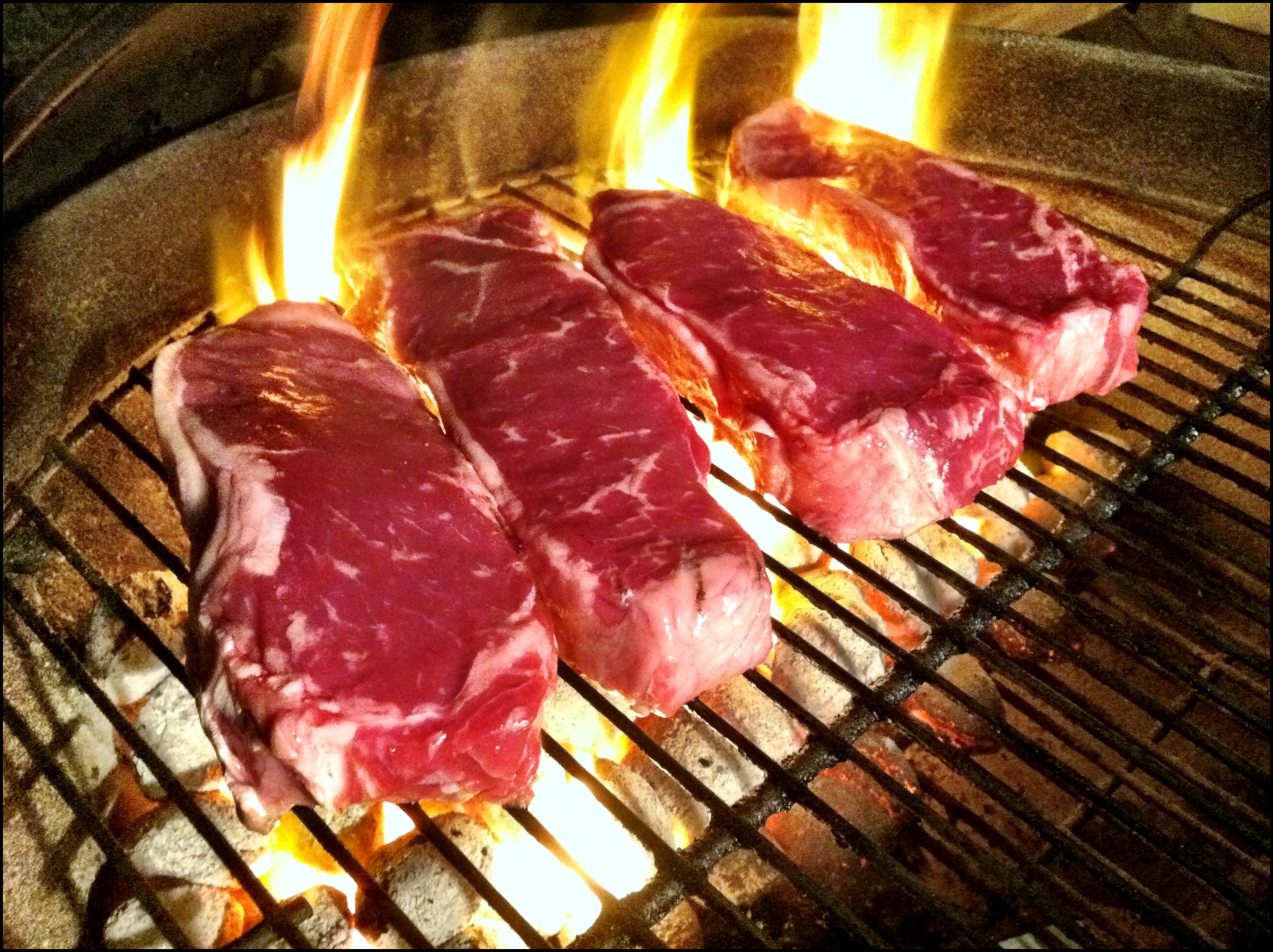 Four raw marbled steaks searing over open flames on a charcoal grill