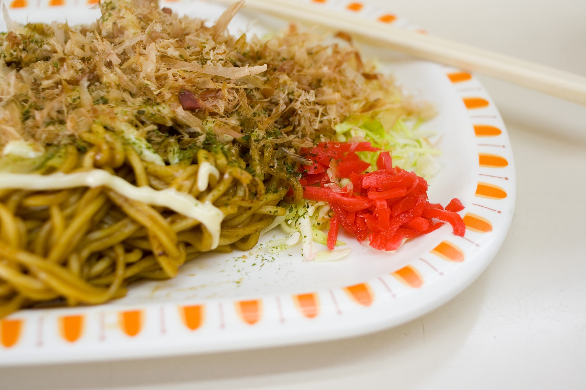 A plate of fried noodles topped with bonito flakes and pickled ginger, with chopsticks resting on the plate