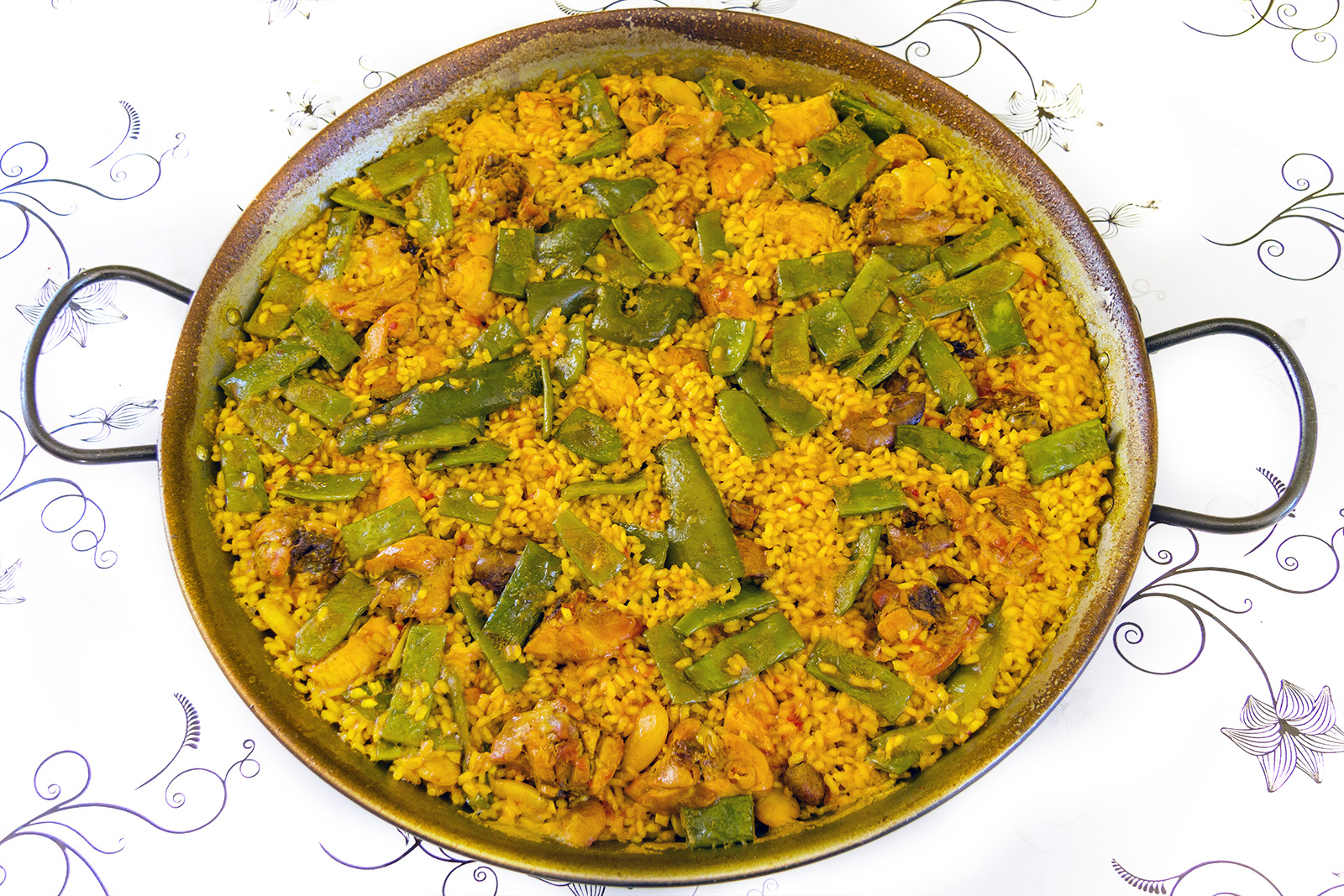 A wide pan of Valencian paella with green beans and chicken on a patterned tablecloth