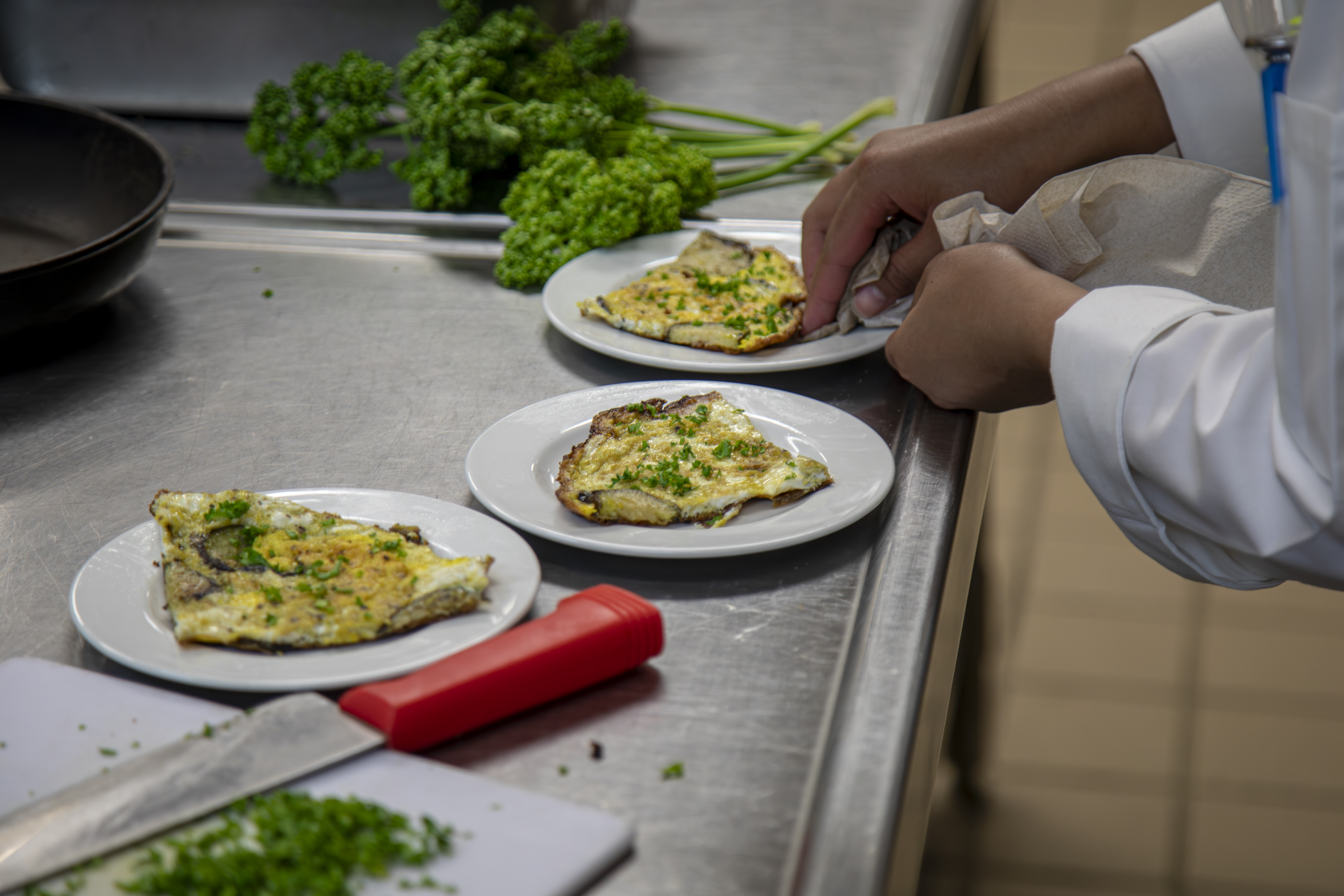 A cook's hands finishing plates of herb omelette on a stainless steel kitchen counter