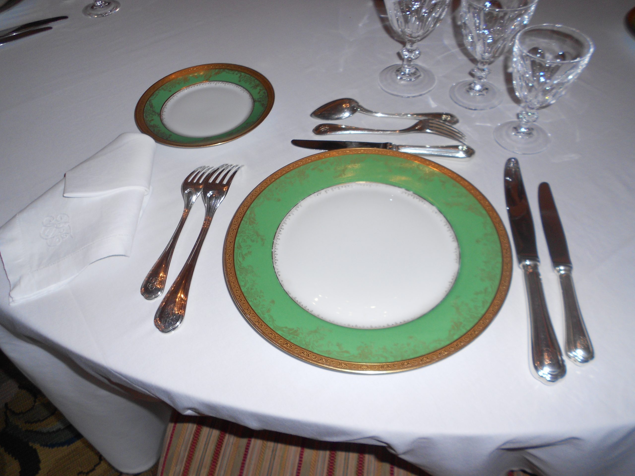 A formal place setting with gilt-rimmed plates, silver cutlery and crystal glasses on a white tablecloth