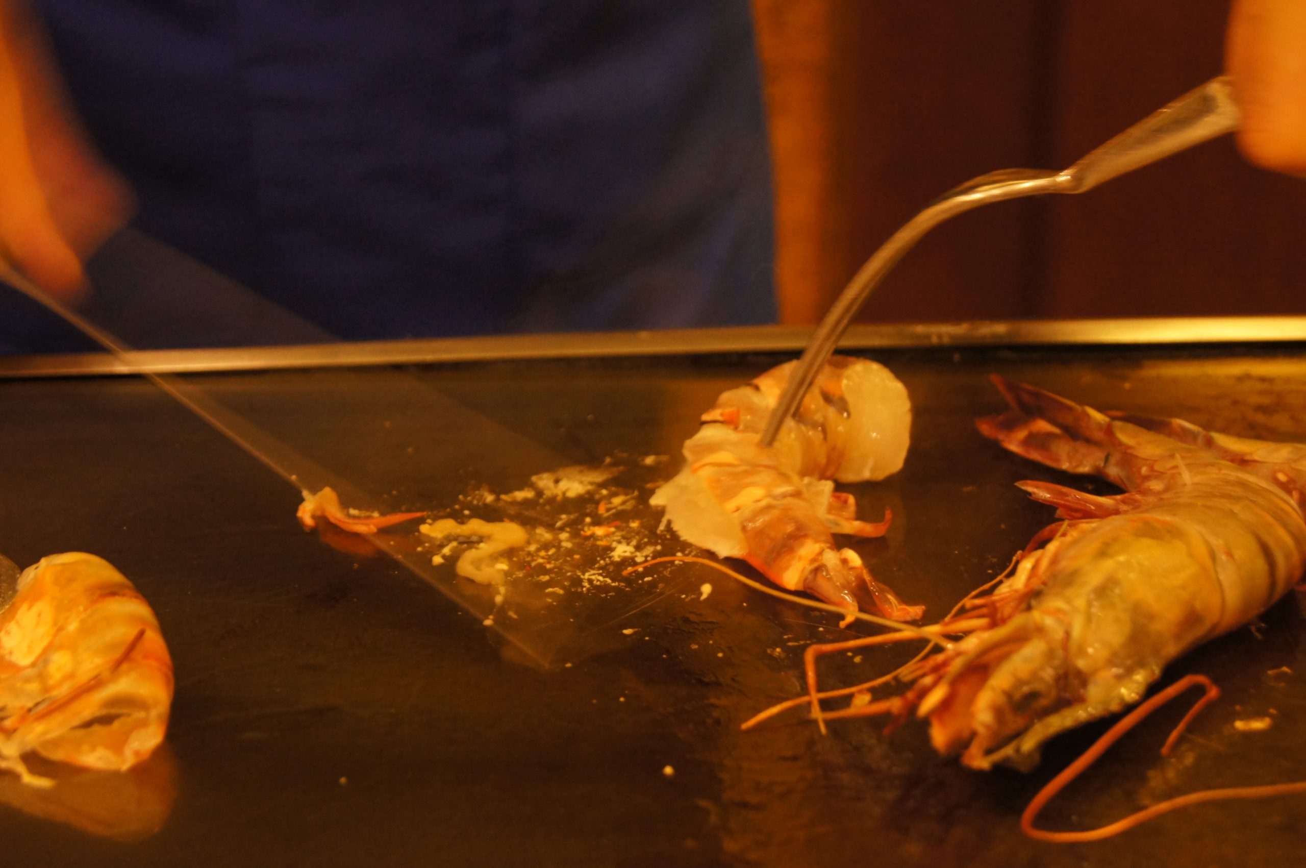 A chef cooking king prawns on a flat griddle directly in front of diners, using a long knife and fork