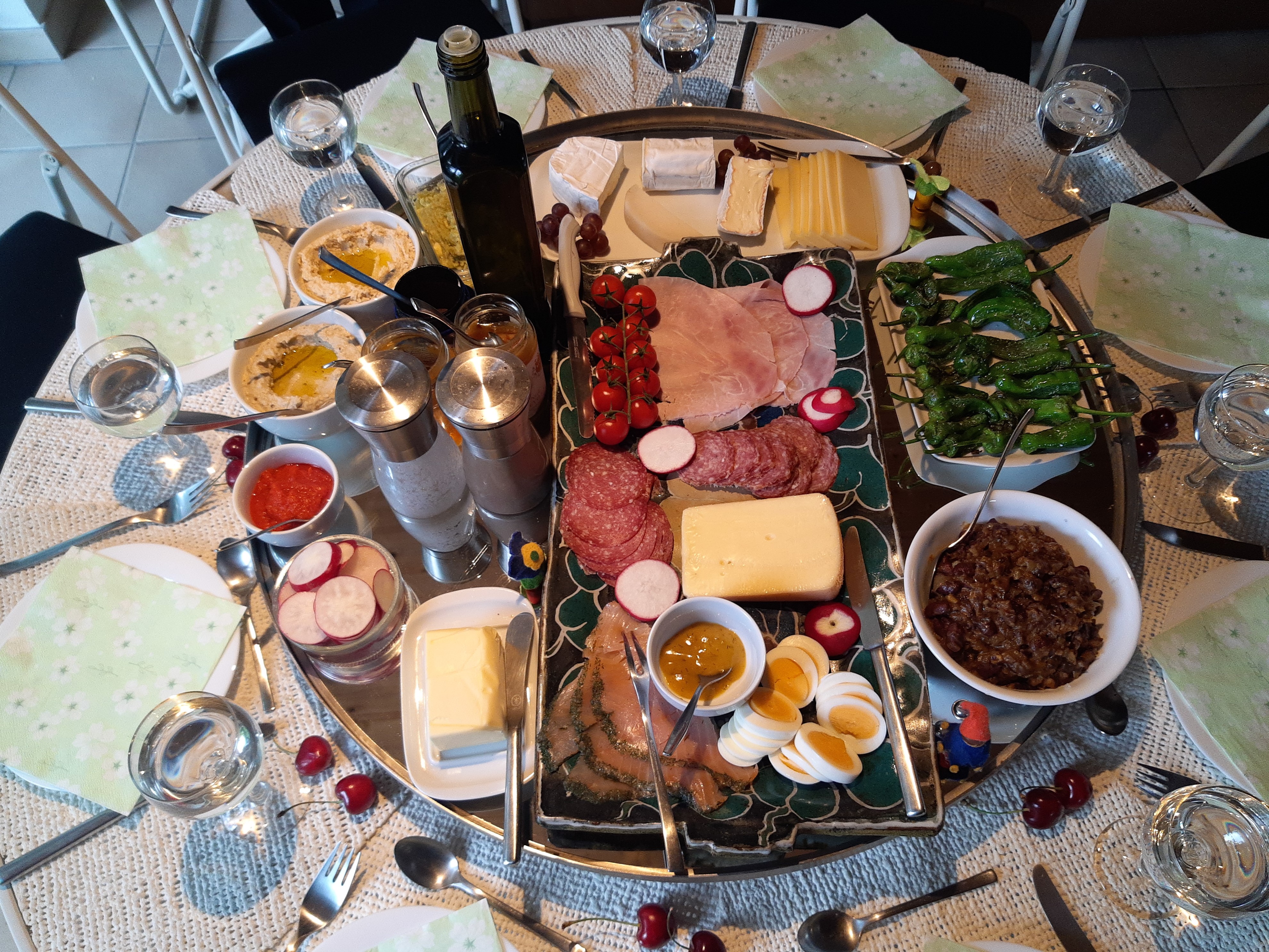 A round table set for brunch with cured meats, cheeses, sliced eggs, peppers and dips on a central platter