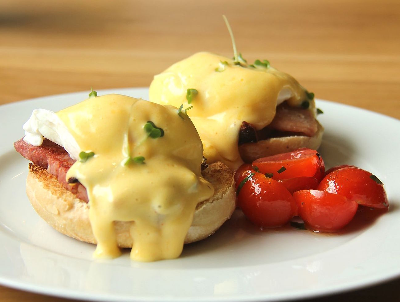 Two eggs Benedict topped with hollandaise and chives beside roasted cherry tomatoes on a white plate
